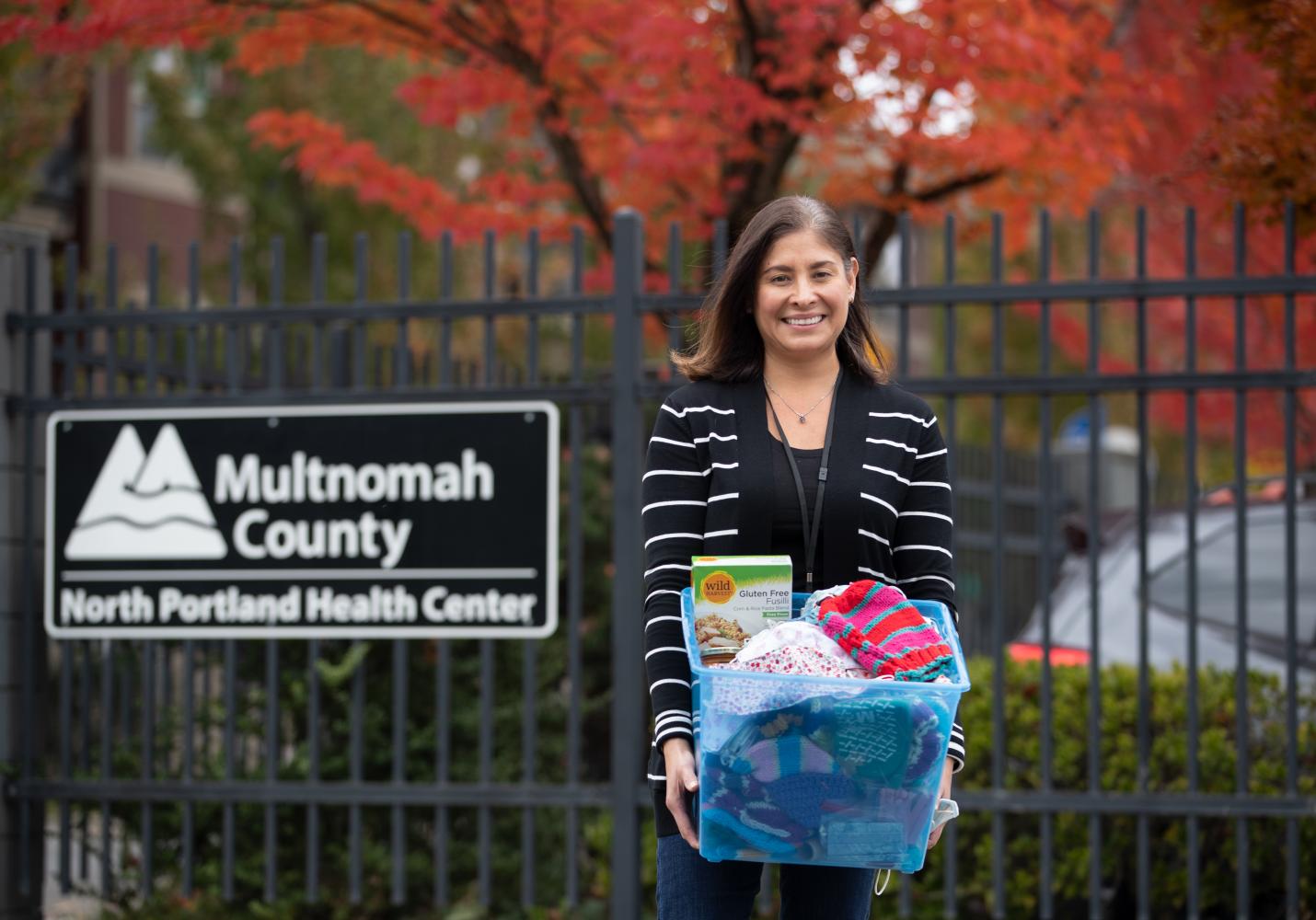 A woman stands near a North Portland Health Center sign and is holding a box full of food and clothing.