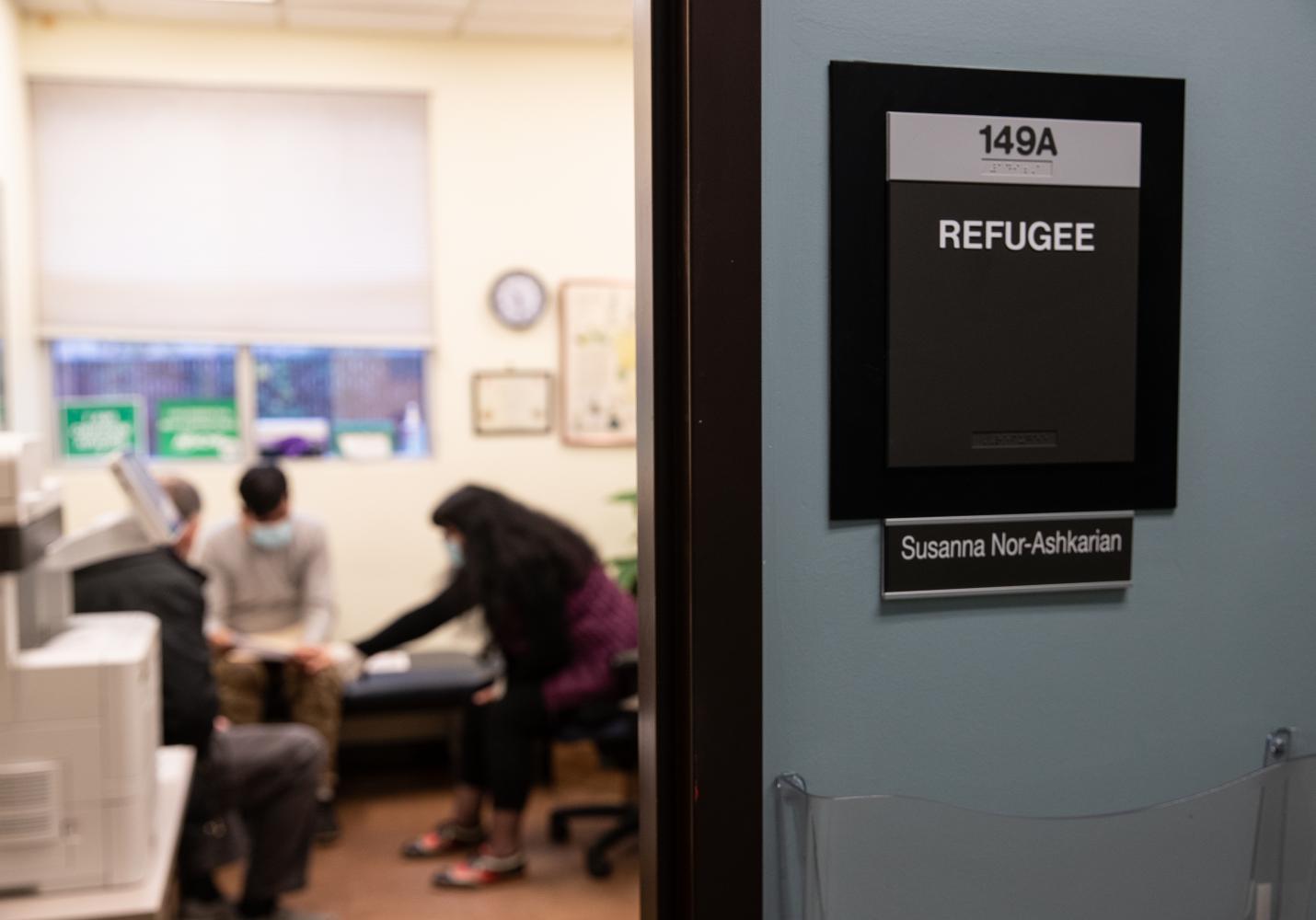 A health care provider works with a family in a room with a sign outside that reads Refugee.