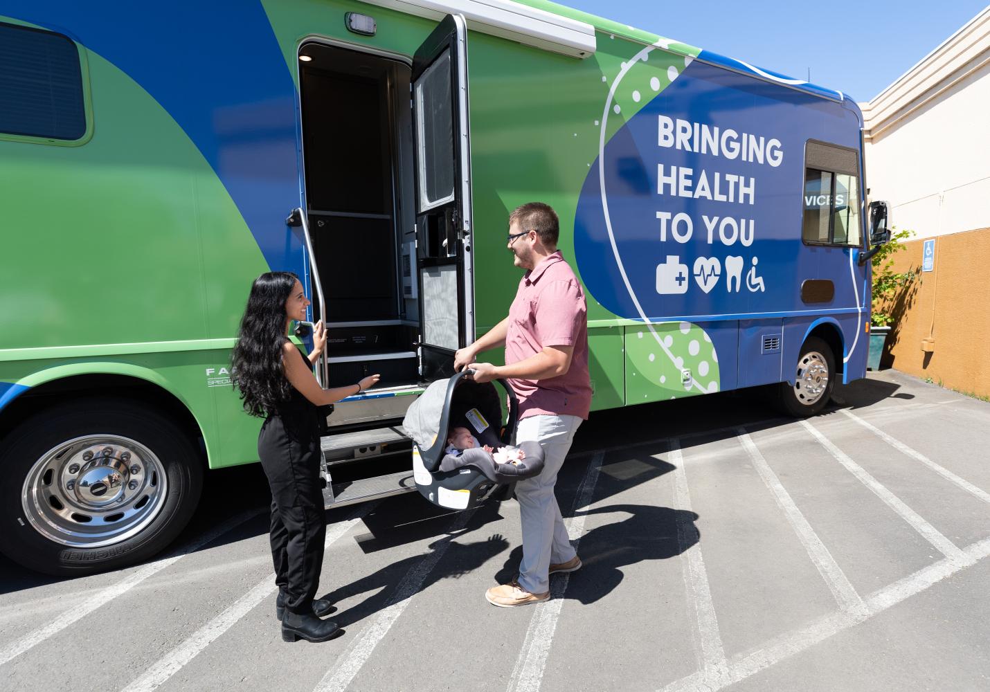 A mobile clinic team stands outside of a mobile clinic medical van with a man who is holding a car seat with a baby in it.