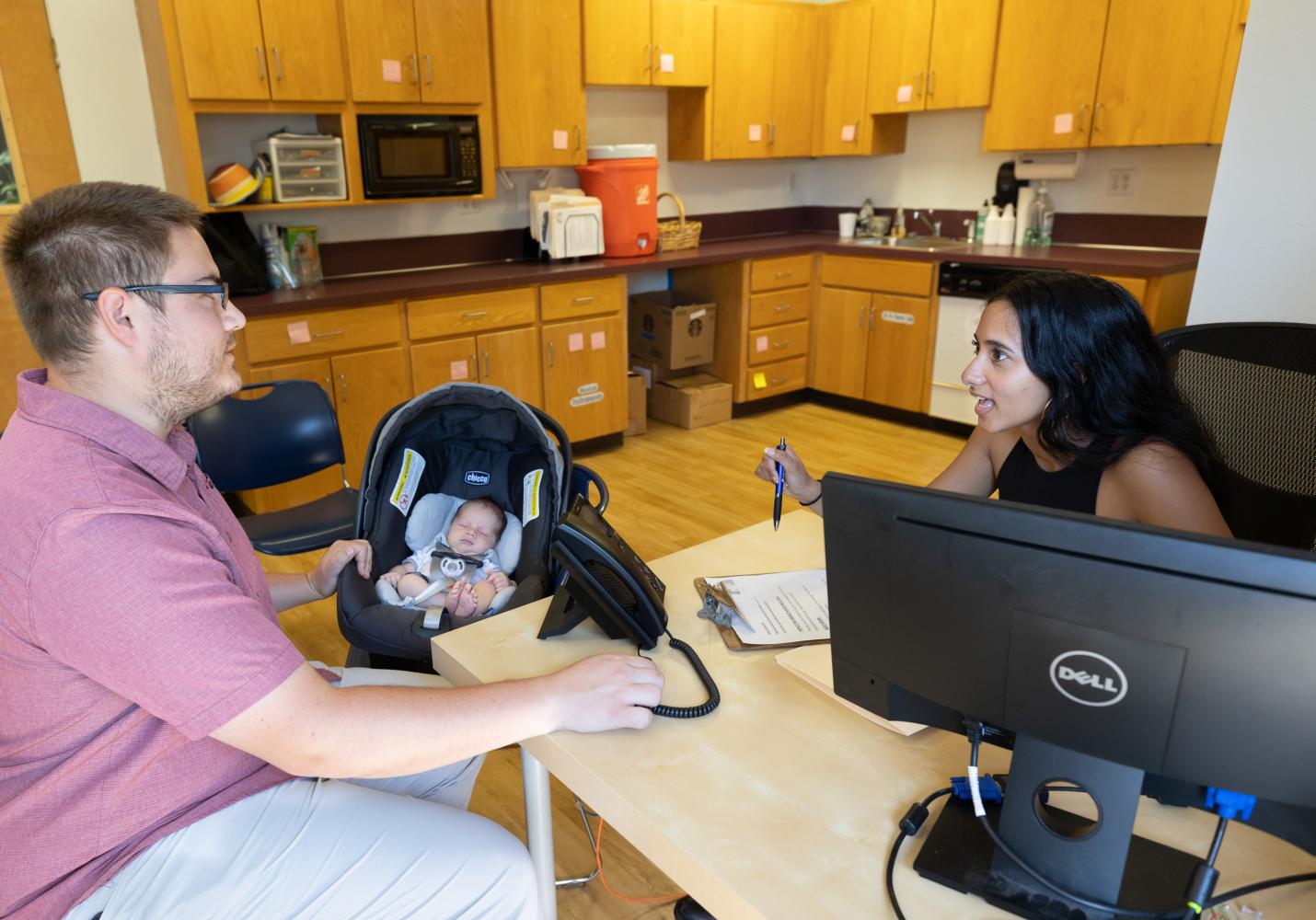 A health care team member talks with a patient while a baby sleeps in a car seat next to them.