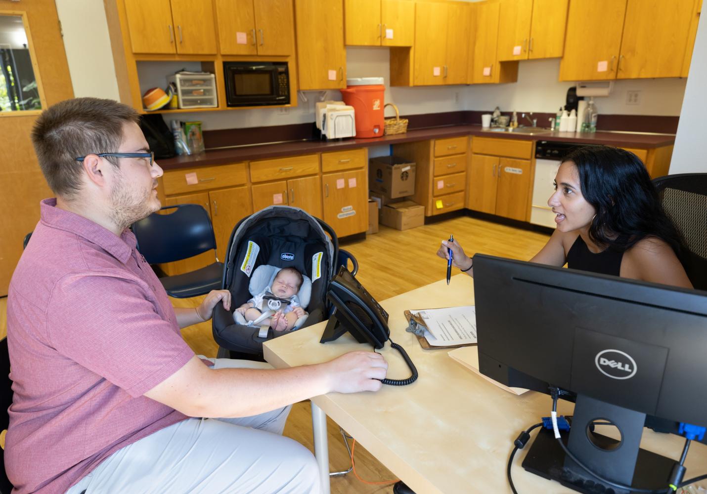 A health care team member talks with a patient while a baby sleeps in a car seat next to them.