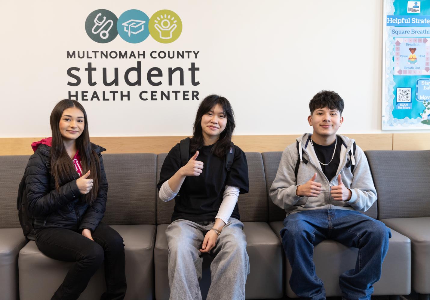 Three teenagers sit together in a Student Health Center waiting area while giving thumbs up.