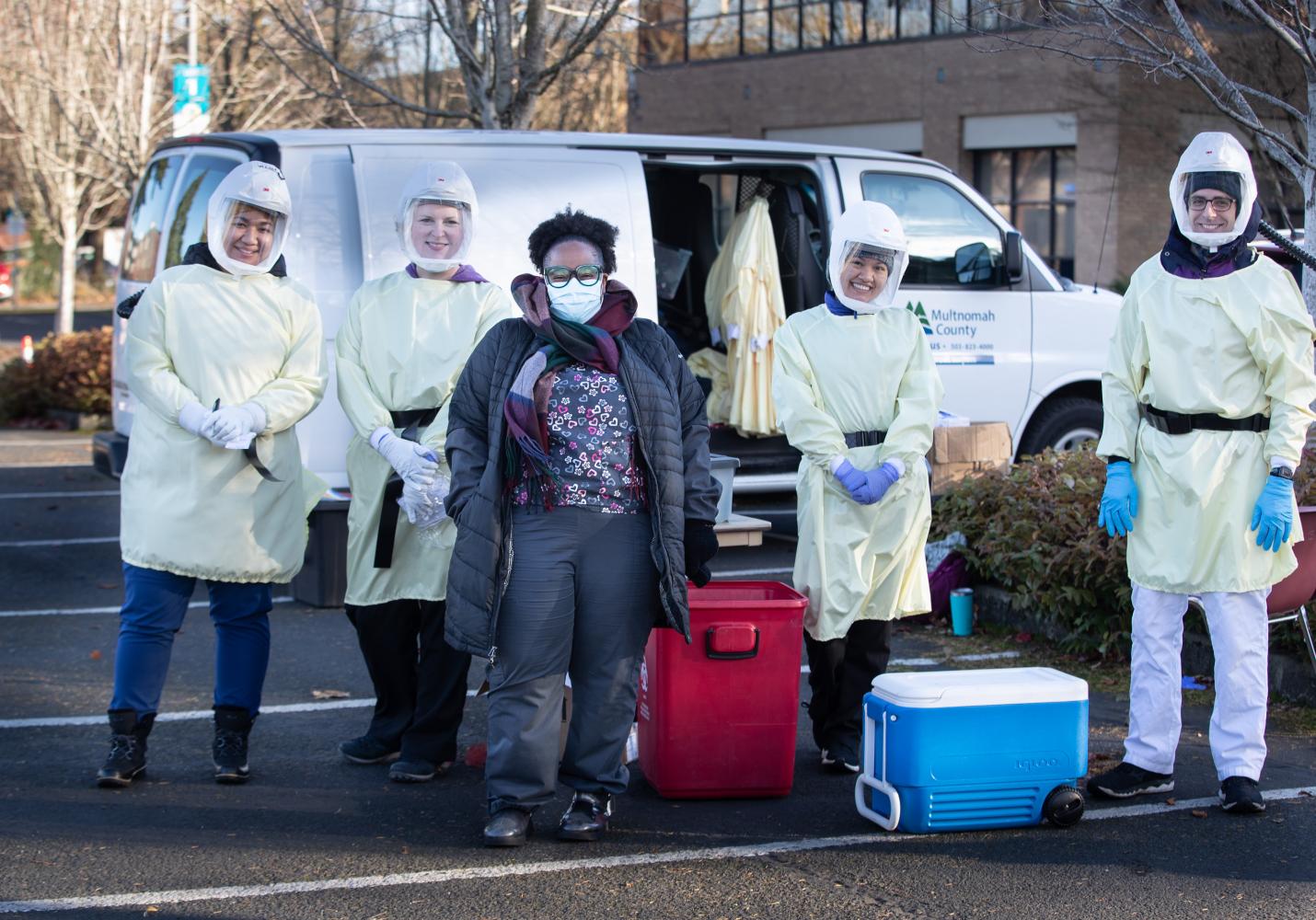 Health care team members wearing protective gear stand together in front of a van.