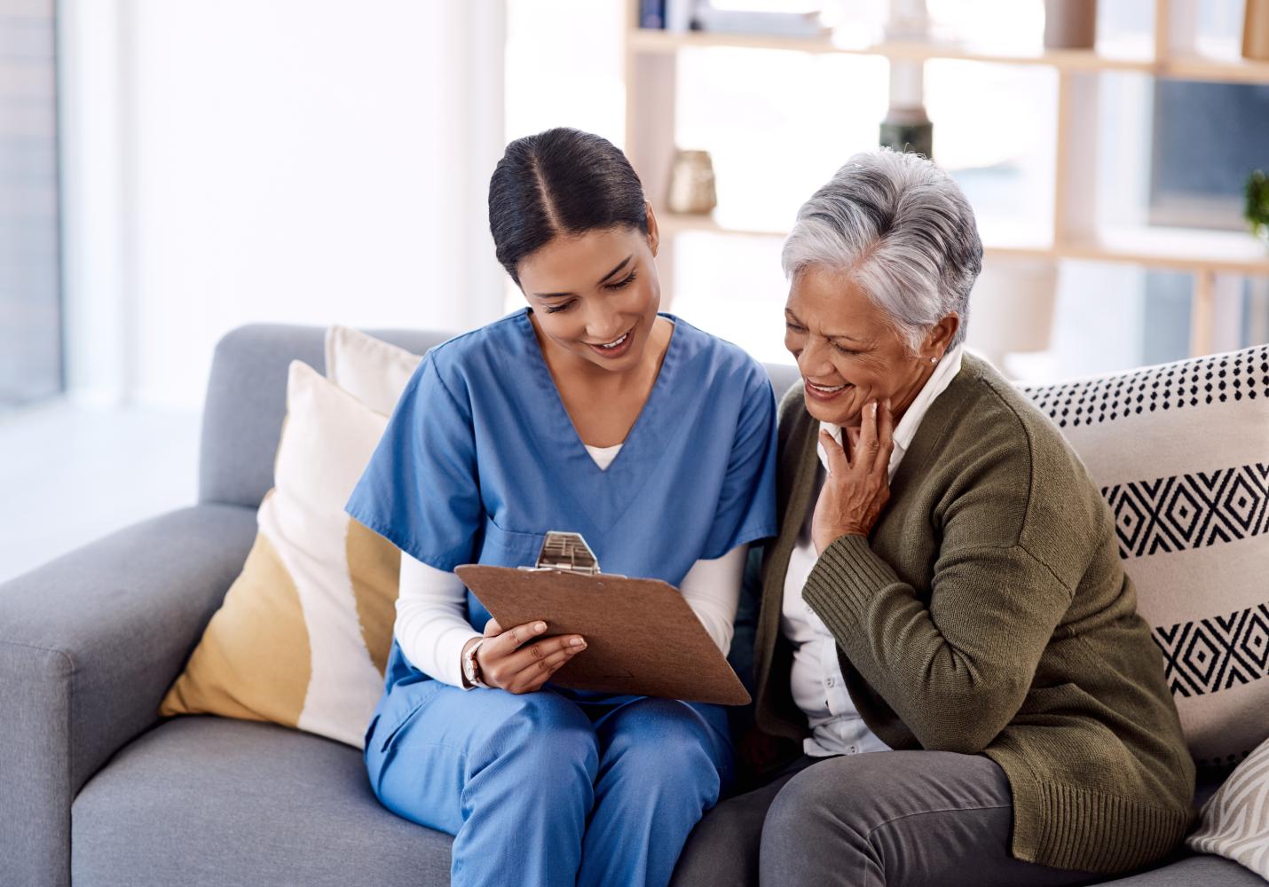 A nurse in blue scrubs reads off paperwork from a clipboard to an elder woman patient