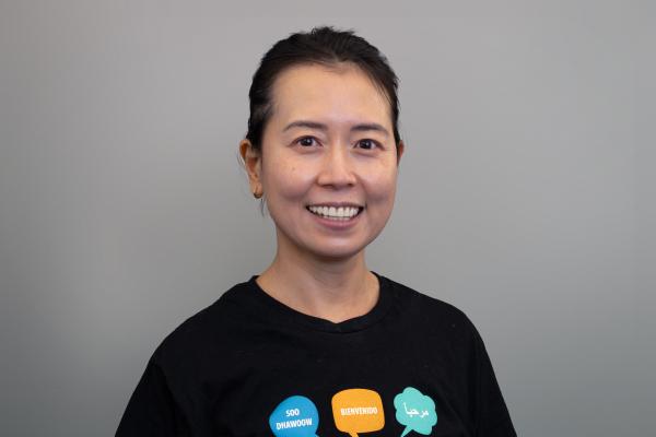 Mya Tran is dressed in a black t-shirt with illustrations on and smiling in front of a grey backdrop